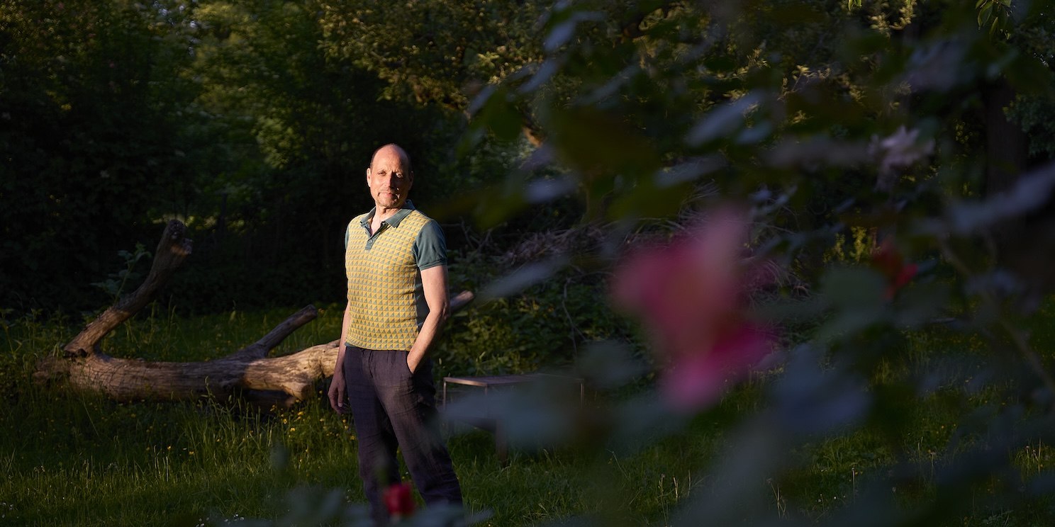 Georg Lolos steht in einem Shirt auf einer Wiese, umgeben von dunklem Grün.