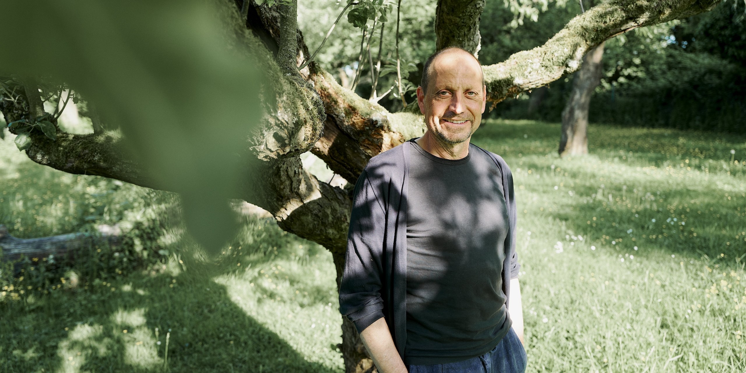 Georg steht auf einer grünen Wiese vor einem Baum. Er lächelt und trägt ein schwarzes Shirt, über das er eine schwarze, dünne Jacke trägt.