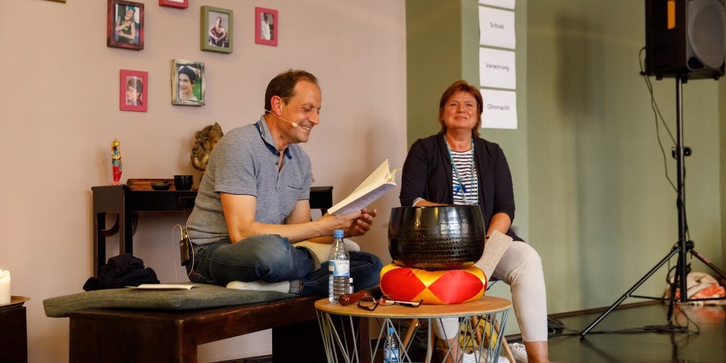 Georg sitzt und liest lächelnd aus einem Buch vor. Neben ihm sitzt eine Frau und vor ihm steht eine Klangschale auf einem Kissen. Im Hintergrund sind viele verschiedene Fotos zu sehen.