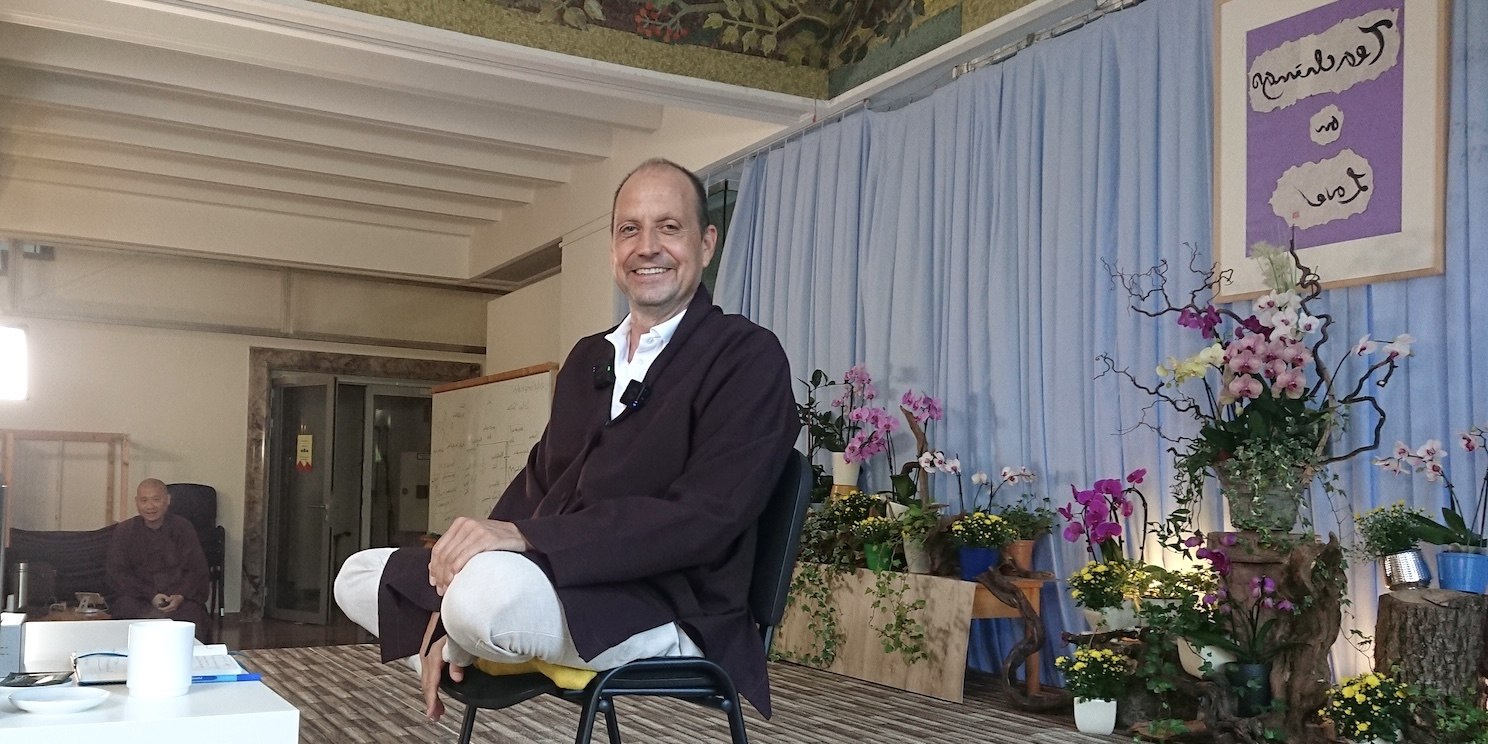 Georg sitzt mit Beinen überkreuzt, lächelnd auf einem Stuhl. Hinter ihm im Vortragsraum stehen viele Orchideen.