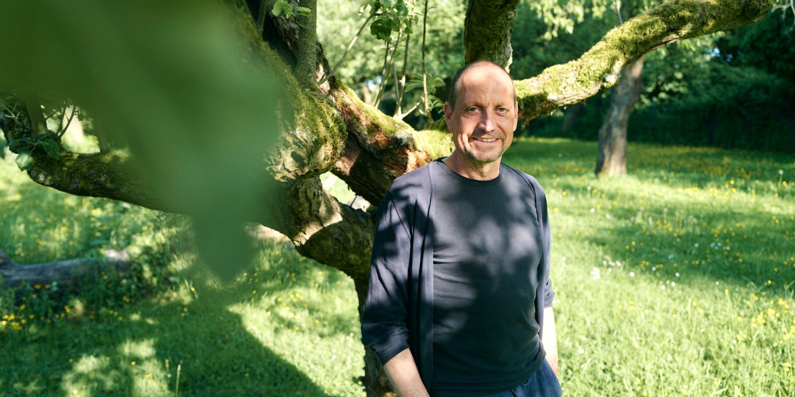 Georg steht auf einer grünen Wiese vor einem Baum. Er lächelt und trägt ein schwarzes Shirt, über das er eine schwarze, dünne Jacke trägt.